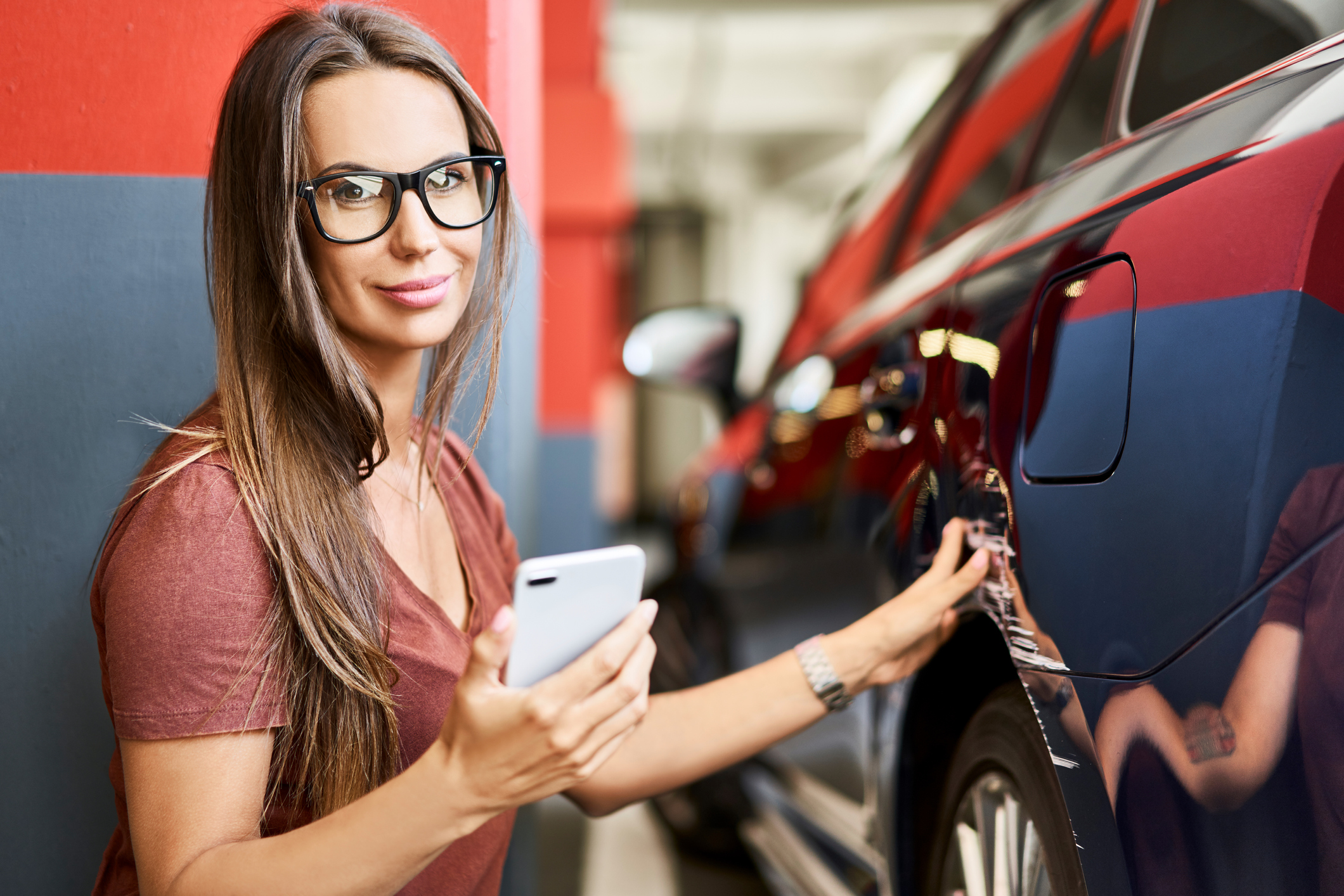 A person looks at a scratch on a car while holding a cellphone.