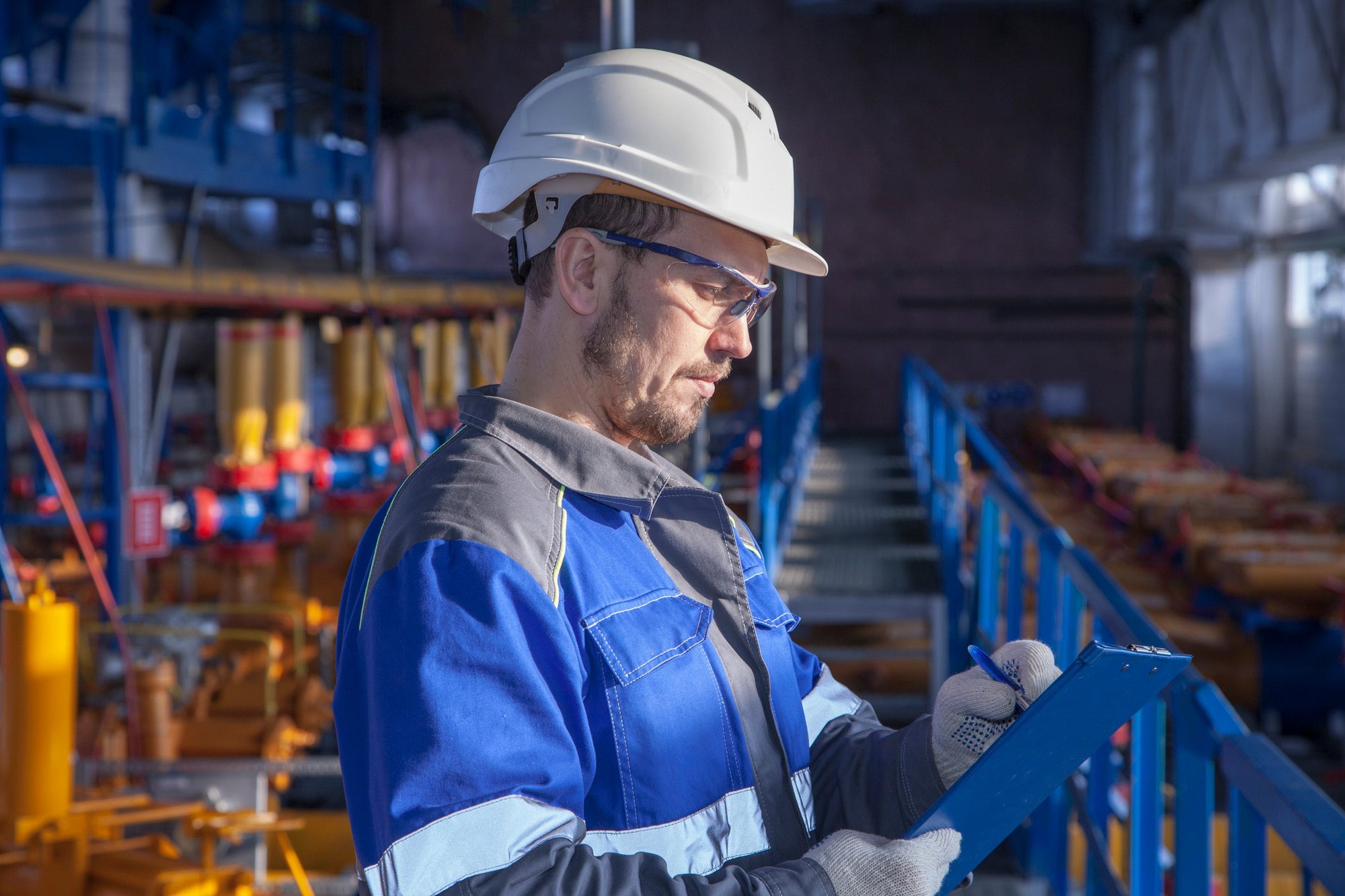 An operator working in an industrial setting wearing a hardhat writes on a clipboard.