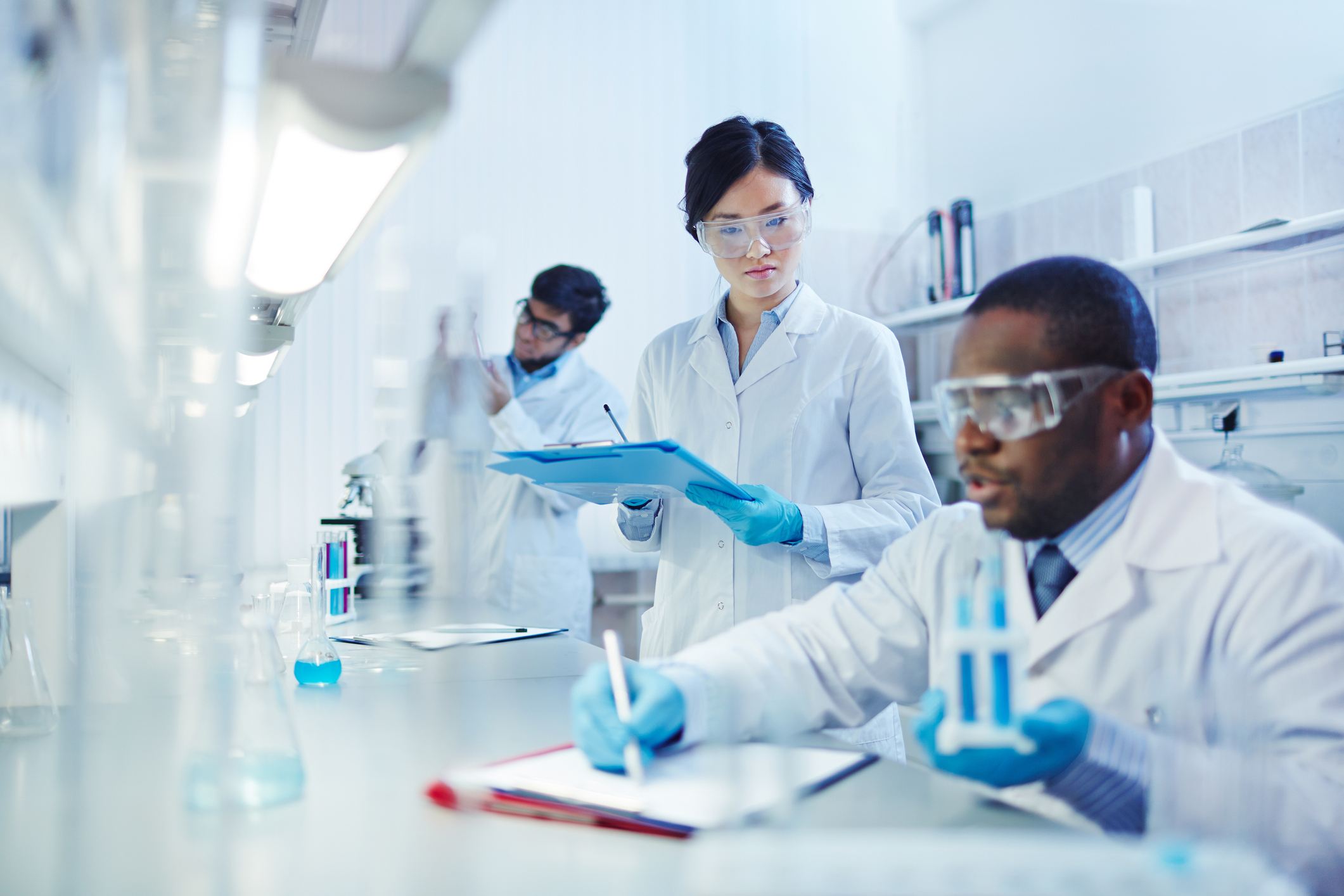 Three scientific researchers take notes in a lab while wearing gloves and goggles.
