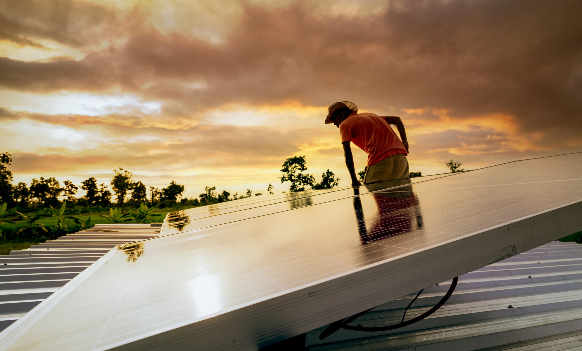 Person lowers a solar panel on to a roof at sunset.