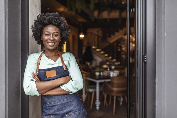 Person with arms crossed, wearing an apron and standing outside a shop.