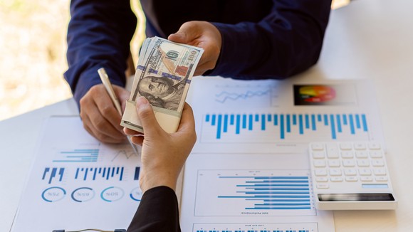 A person hands over a large bundle of cash to another person, over a table full of charts and tables.