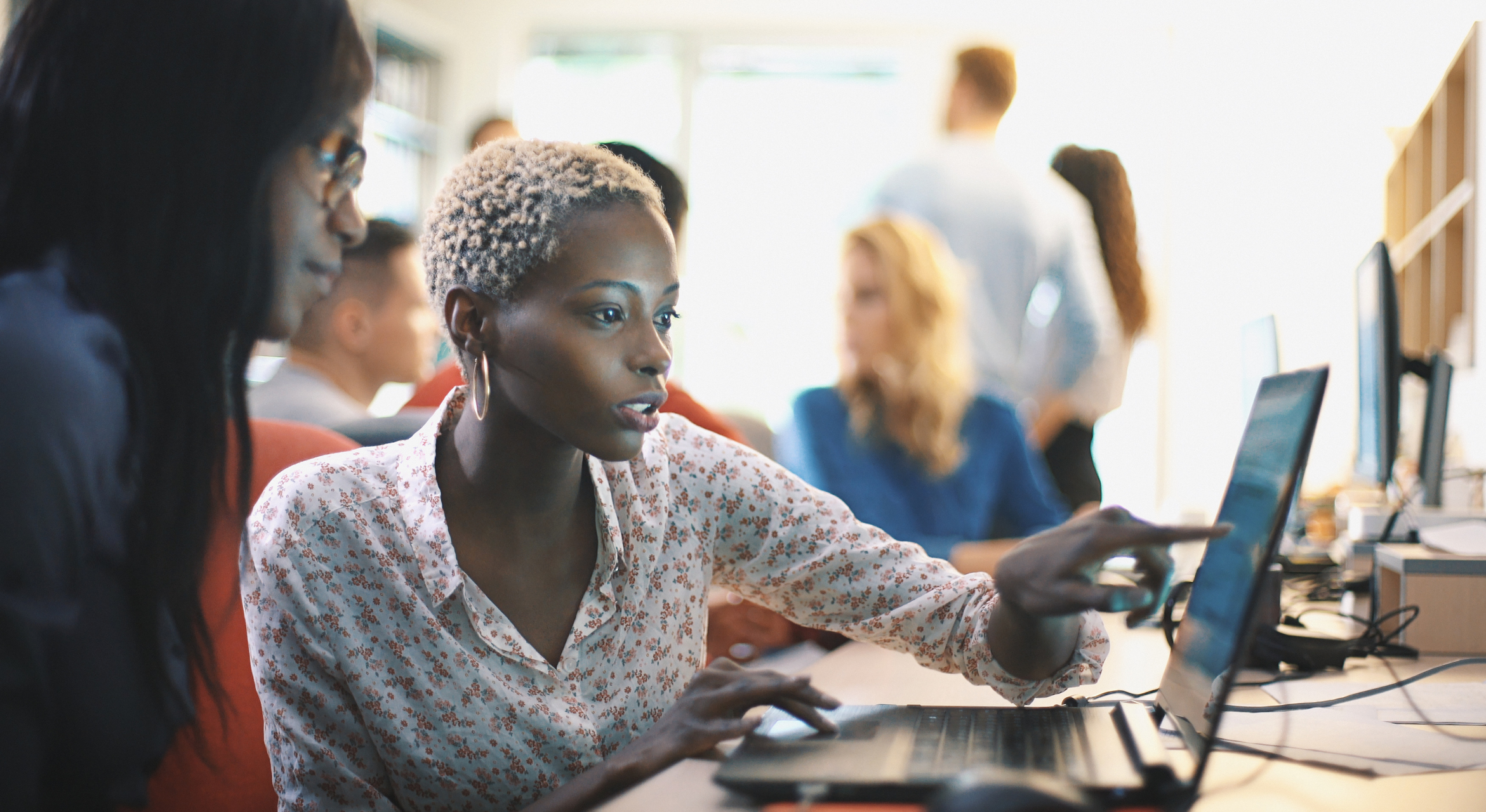 One person points to her computer screen while another person listens.