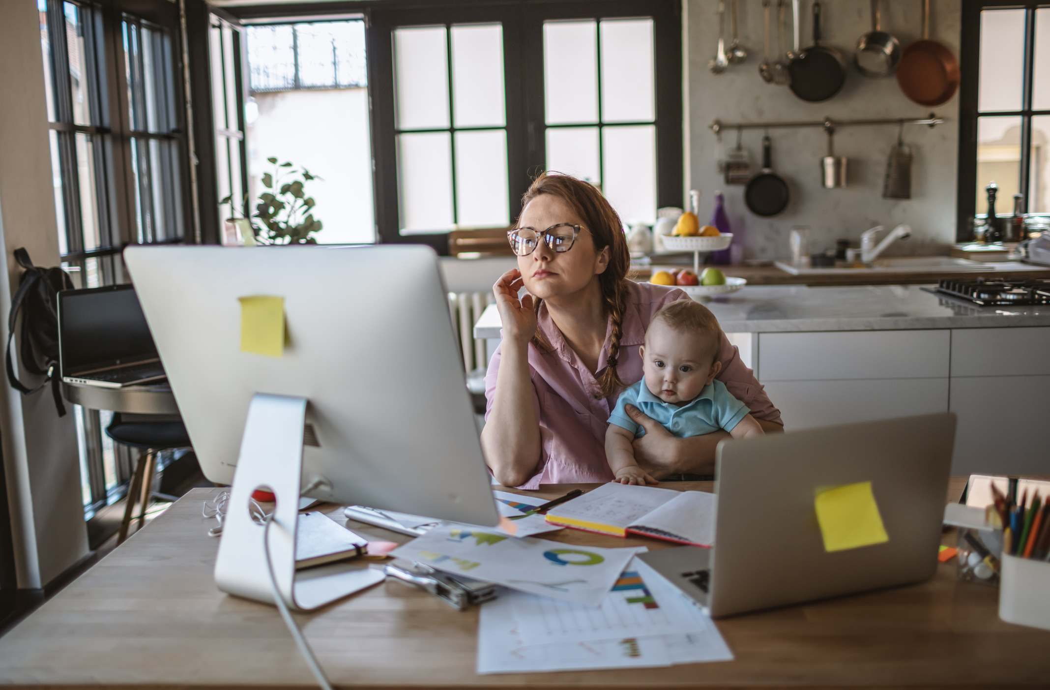 Mother working from home on desktop and laptop while holding a baby.