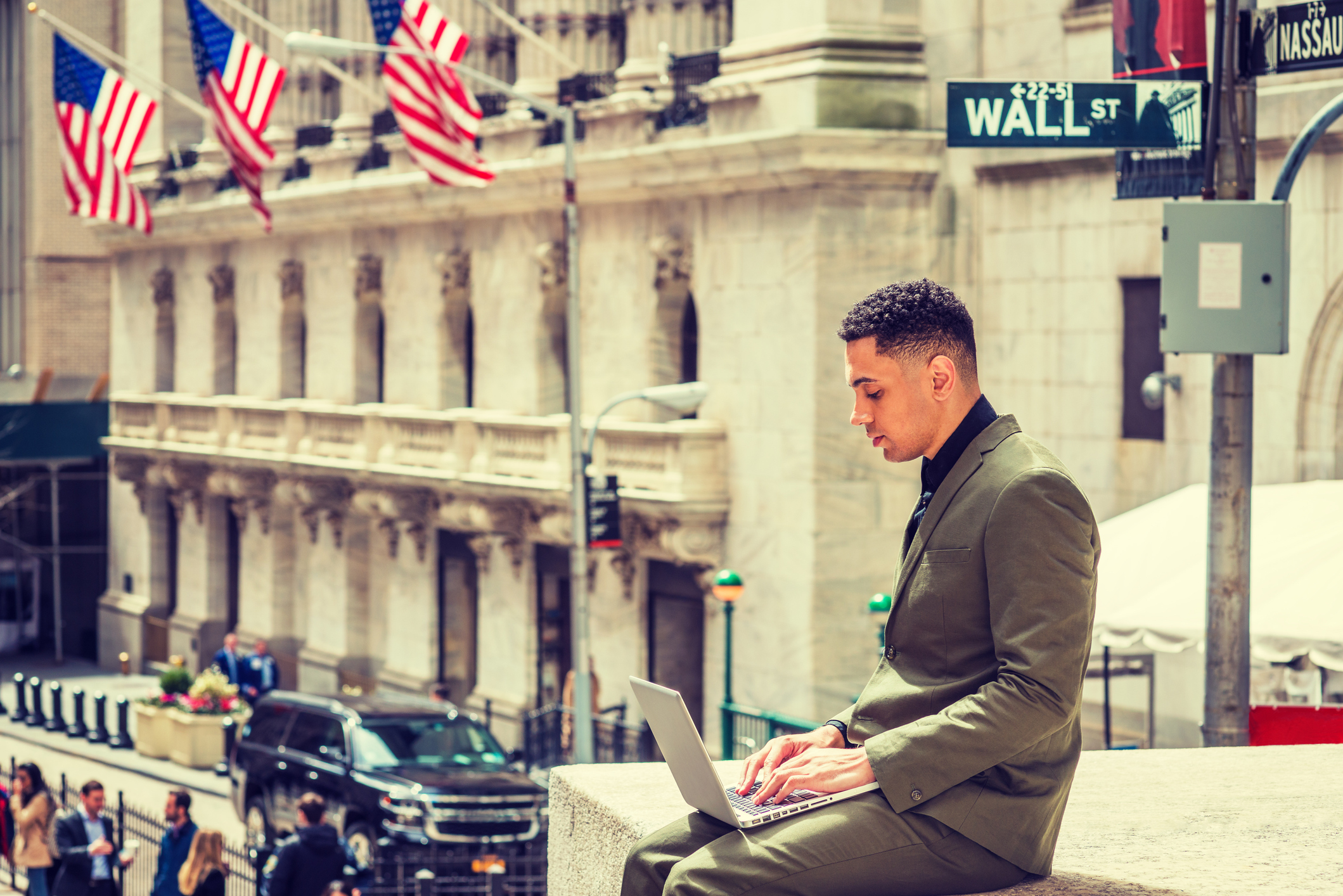 A person working on a laptop with the Wall Street sign and New York Stock Exchange in the background.