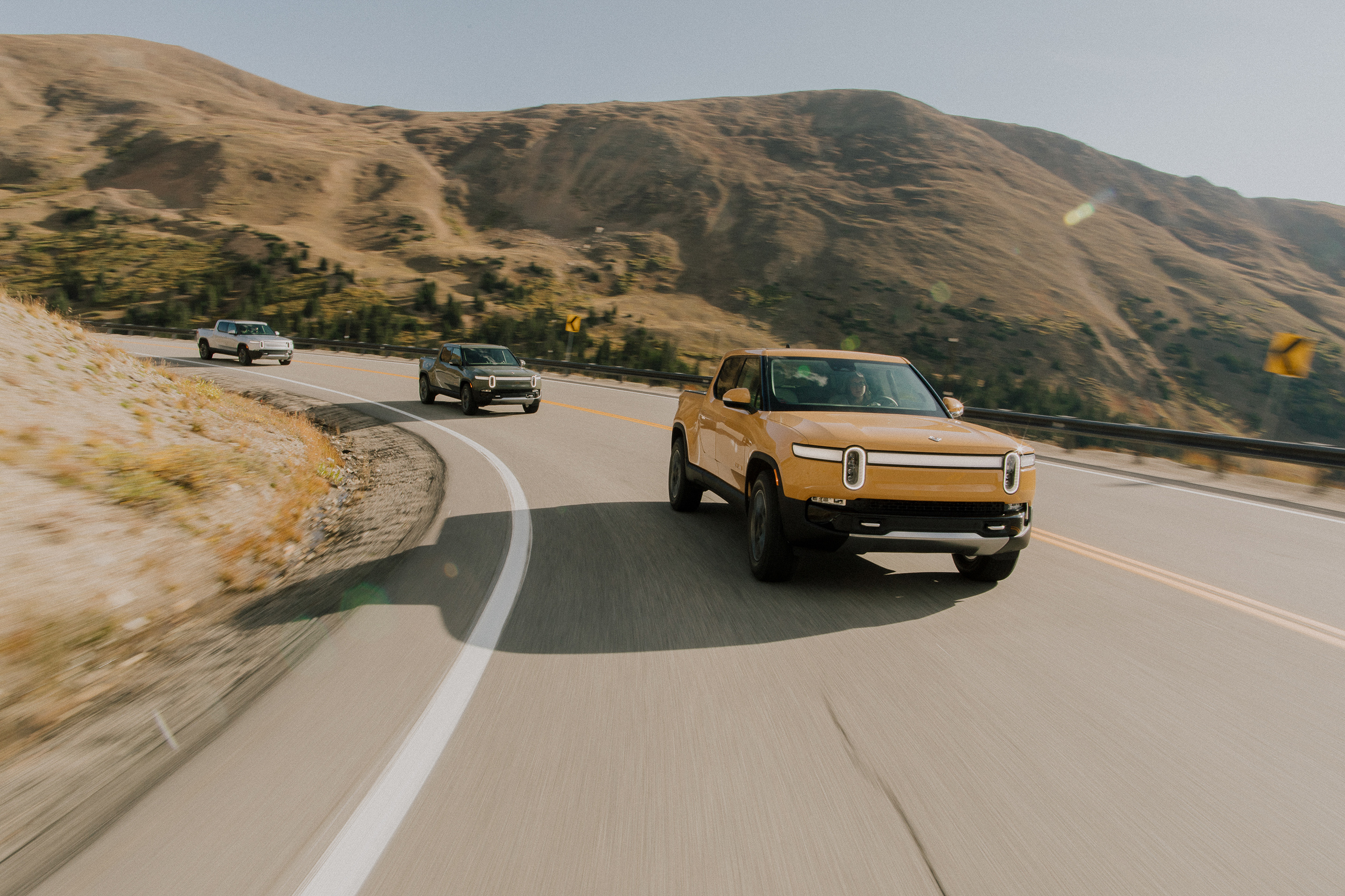 3 Rivian R1T pickup trucks rounding a bend on scenic highway.