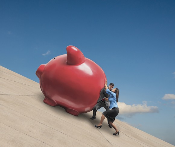 Two people pushing up a huge piggy bank up an incline.