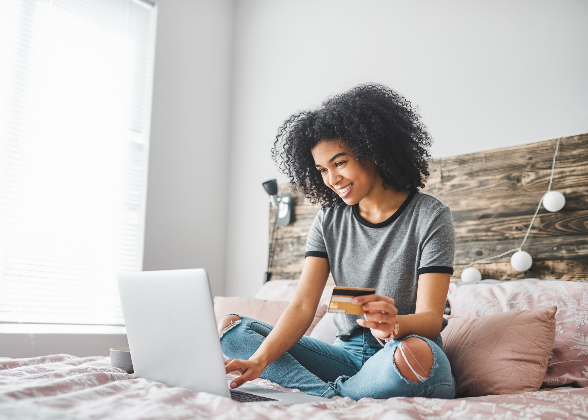 A girl holding a credit card sits on her bed, using her laptop.