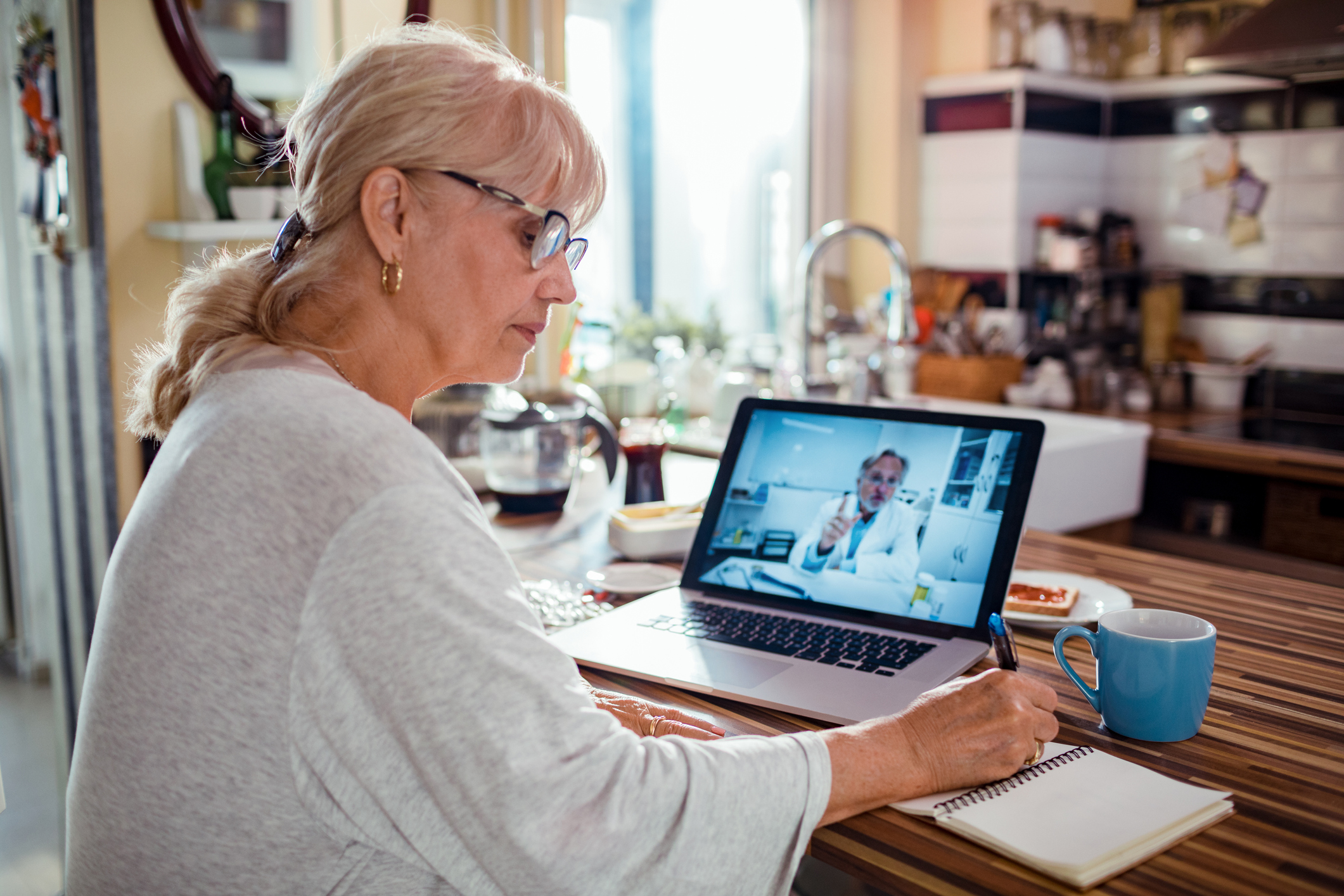A person sits in a kitchen and consults a doctor on a laptop.