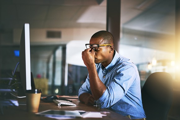 A man pinches the ridge of his nose in agony while sitting at an office desk.