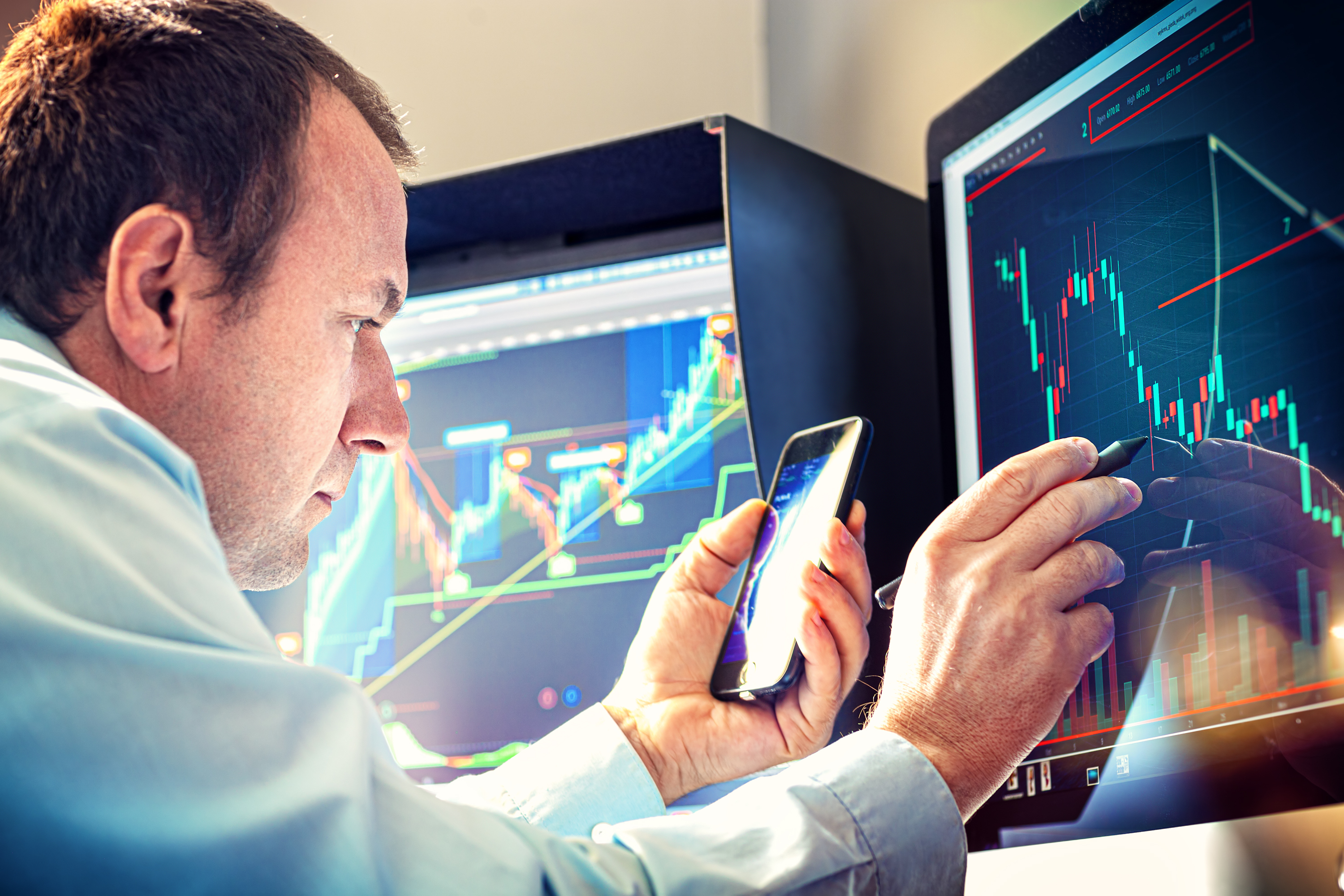 A money manager using a smartphone and stylus to analyze a stock chart on a computer monitor.