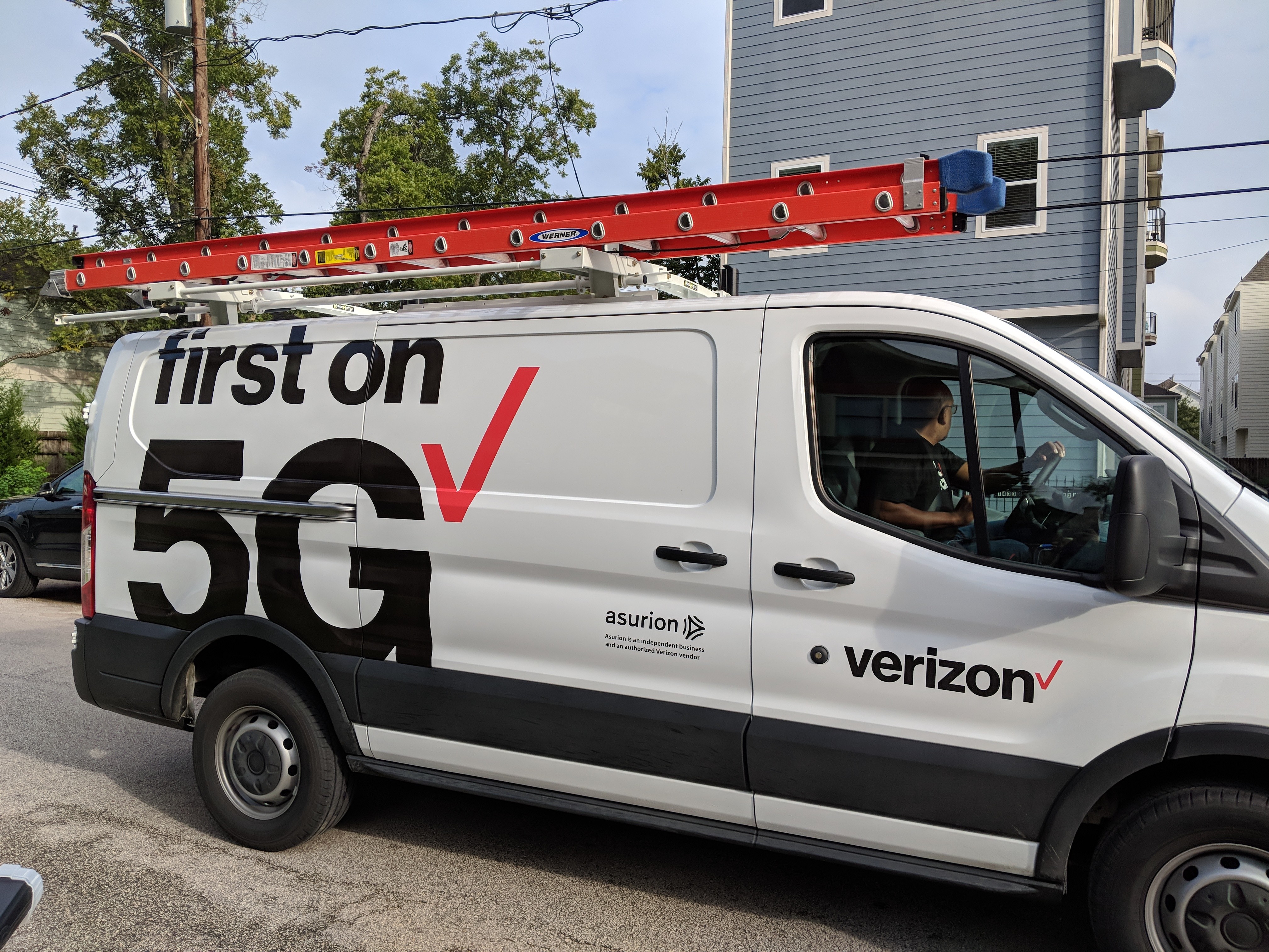 A Verizon maintenance vehicle sits on a roadway near a building. On the side of the van are the words "first on 5G" and a red checkmark.