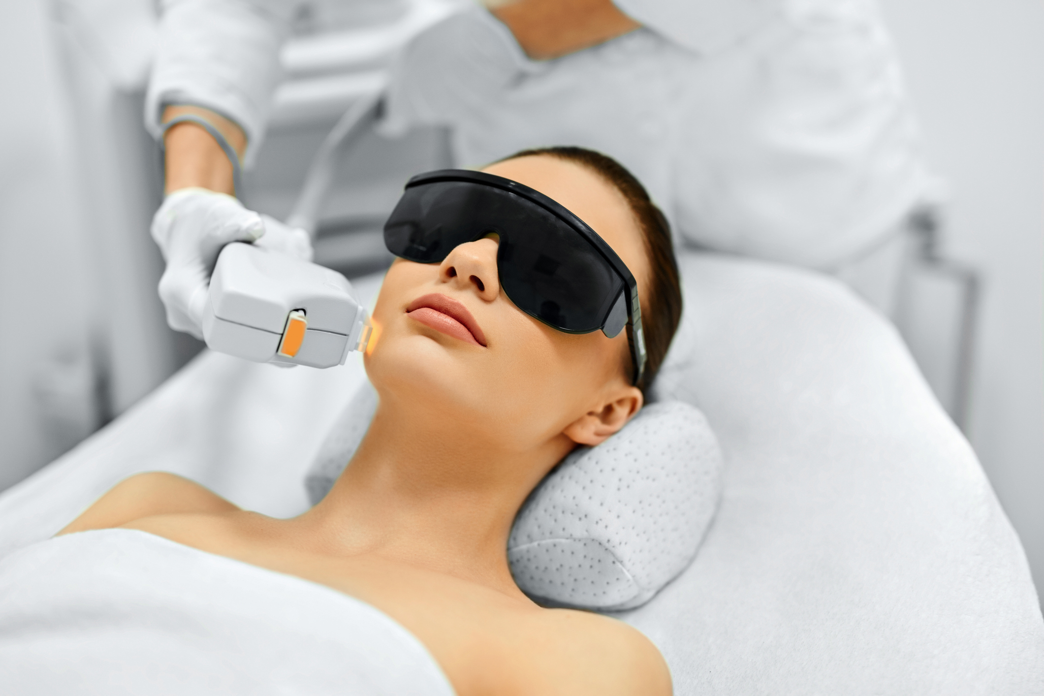 A woman recieves a beautifying skin treatment from a clinician while wearing sunglasses and laying on a bed in a clinic.