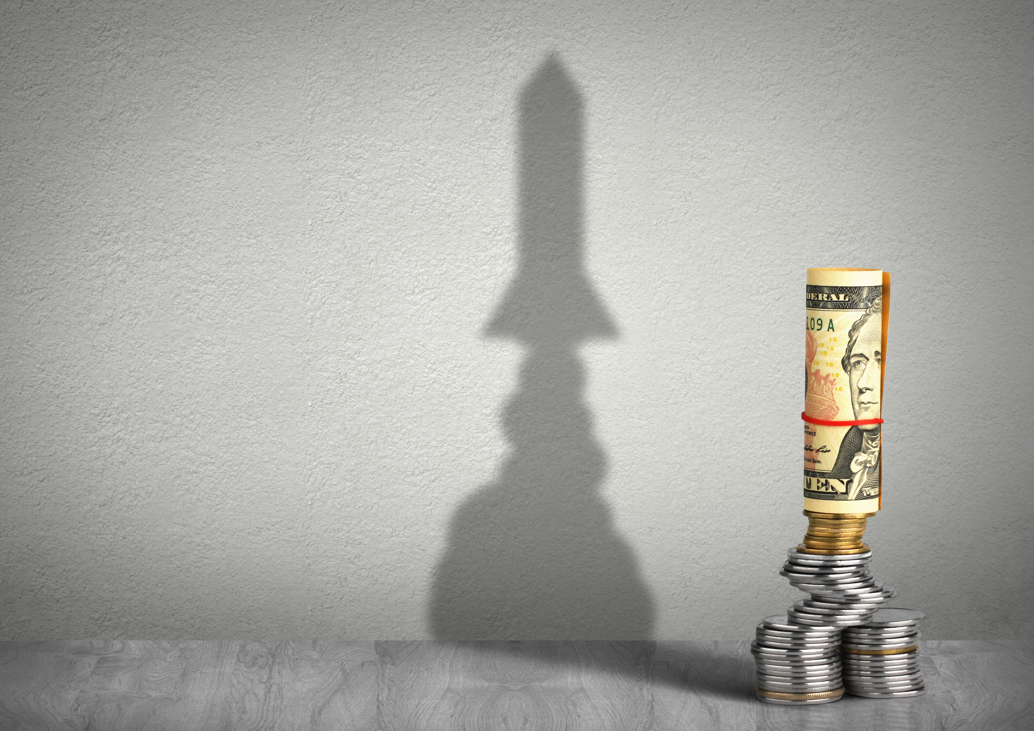 A stack of money casting a shaddow that looks like a rocket ship in the background.