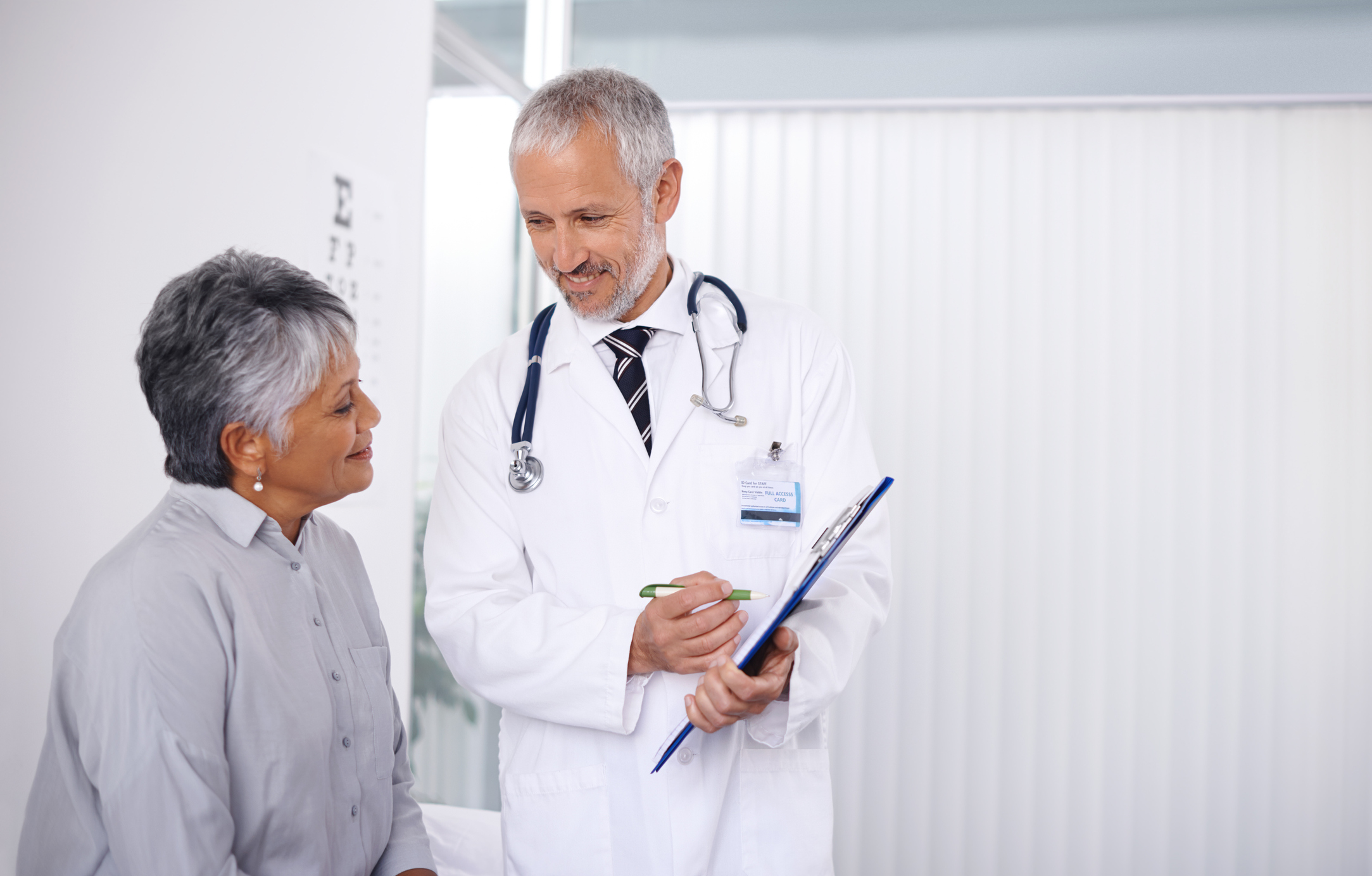 A doctor standing next to a seated patient while holding a clipboard.