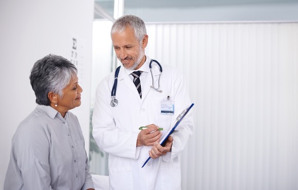 A doctor standing next to a seated patient while holding a clipboard.
