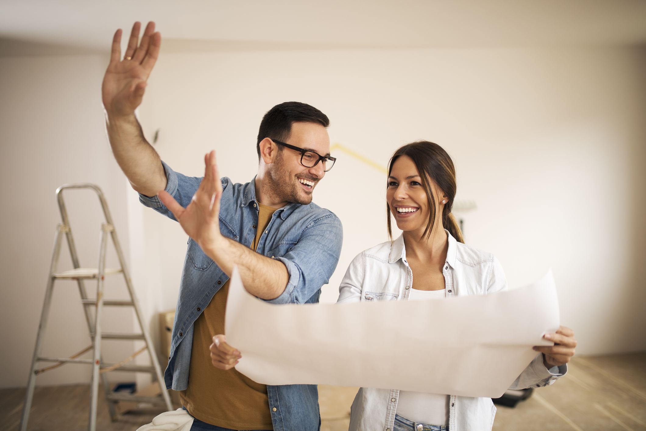 Two people smiling holding remodeling plans with ladder in background.