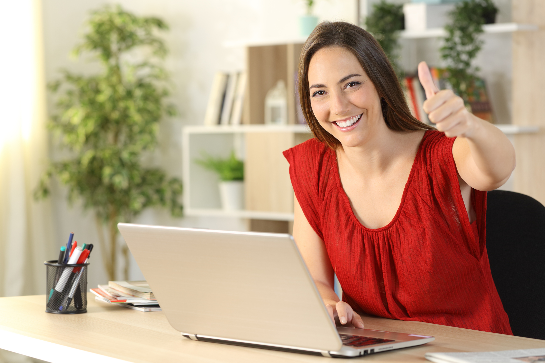 Person in red shirt sitting at laptop in home office smiling and giving thumbs up.