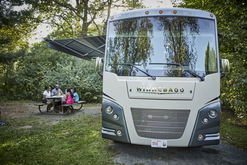 A family eats at a picnic table next to a parked Winnebago RV.