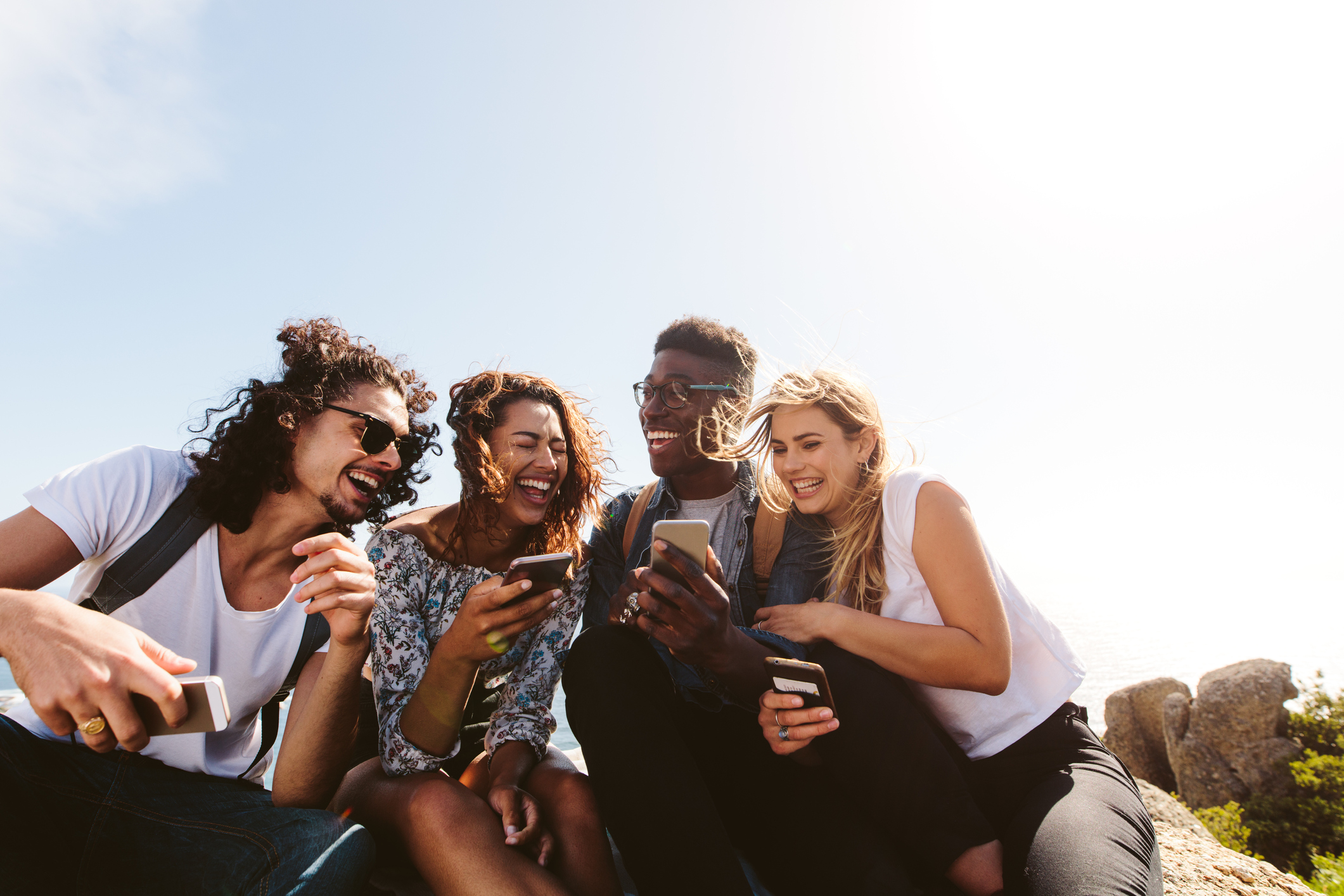 Four young people are laughing while looking at a mobile phone screen.