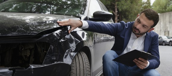 A person with a clipboard reviews damage to a car fender.