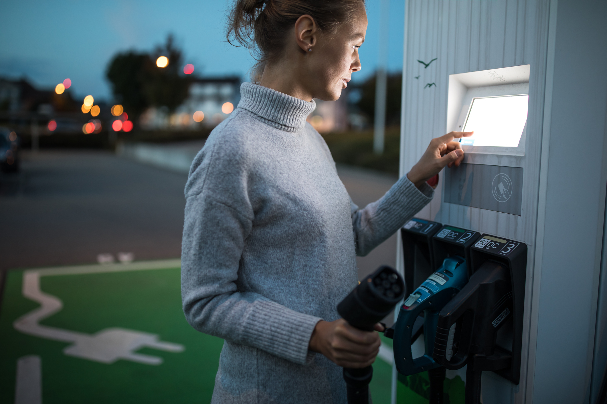 A person holding an electric vehicle charger while punching in the charging station's display board.