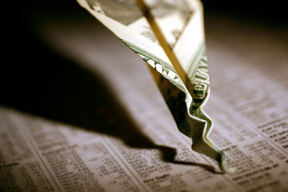 Paper airplane made out of money is crashing into printout of stock tables.