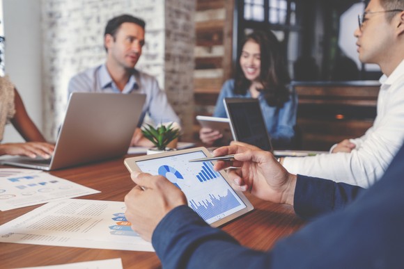 Employees using tablets and laptops to analyze financial metrics during a conference room meeting.