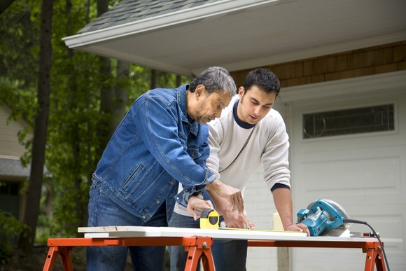 Two men measuring a piece of wood to cut in driveway.