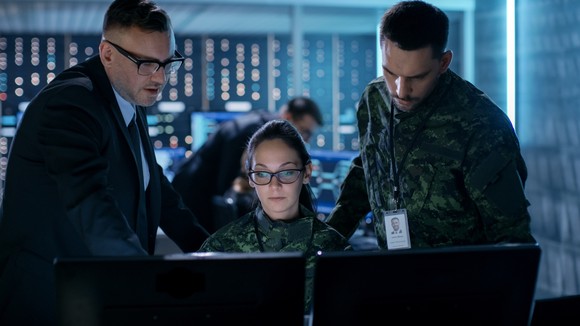 Two soldiers work and an IT professional looking at two monitors.