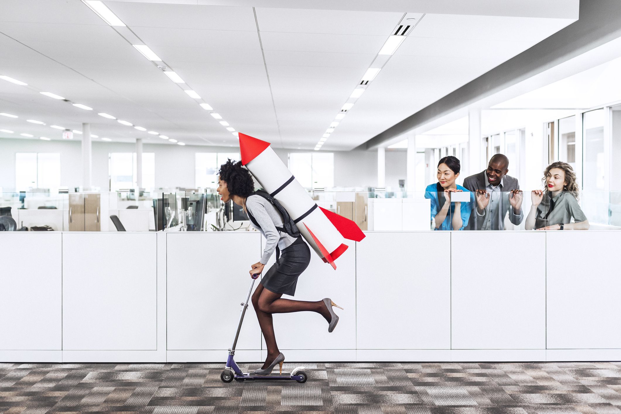 Person on scooter with rocket strapped to their back, rolling through office.