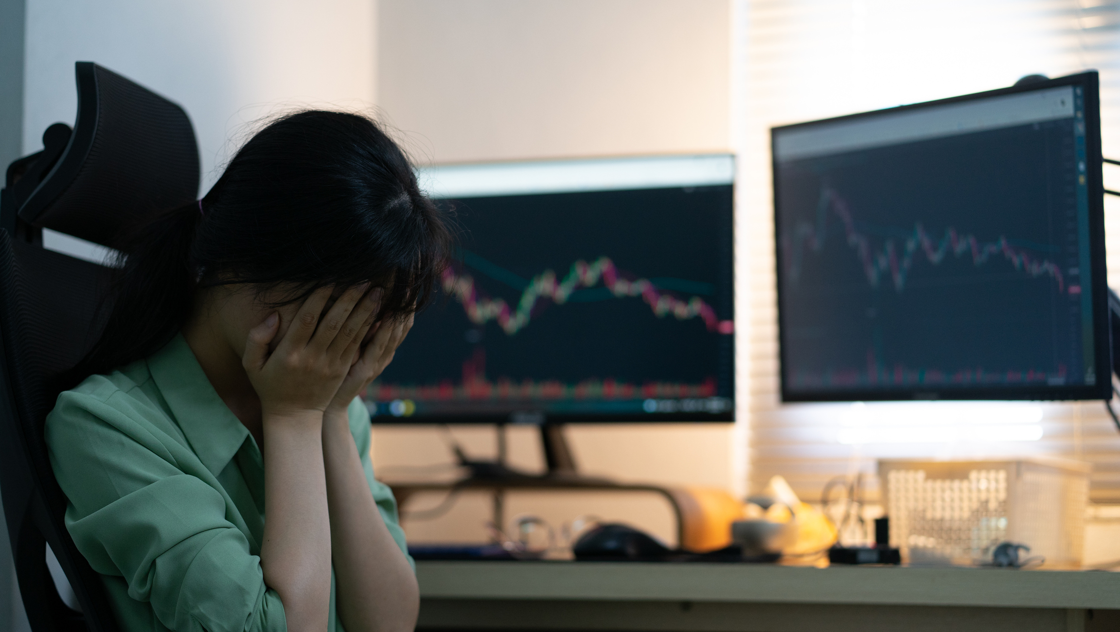 A person holding hands over face with two monitors showing declining stock charts.