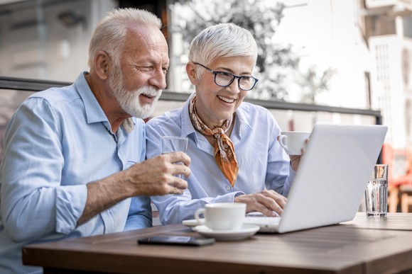 A retired couple smiles as they look at a computer screen while drinking coffee together.