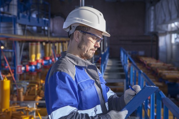 A technician wearing a hard hat and personal protective equipment writes on a clipboard. 