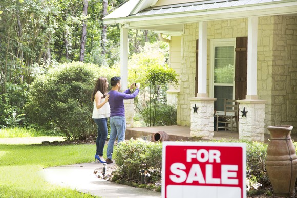 Couple taking a picture of a home with for sale sign in front yard. 