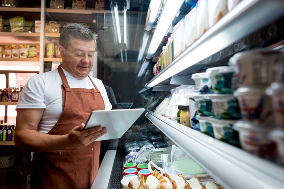 A person standing in a food aisle wearing an apron and holding a tablet.