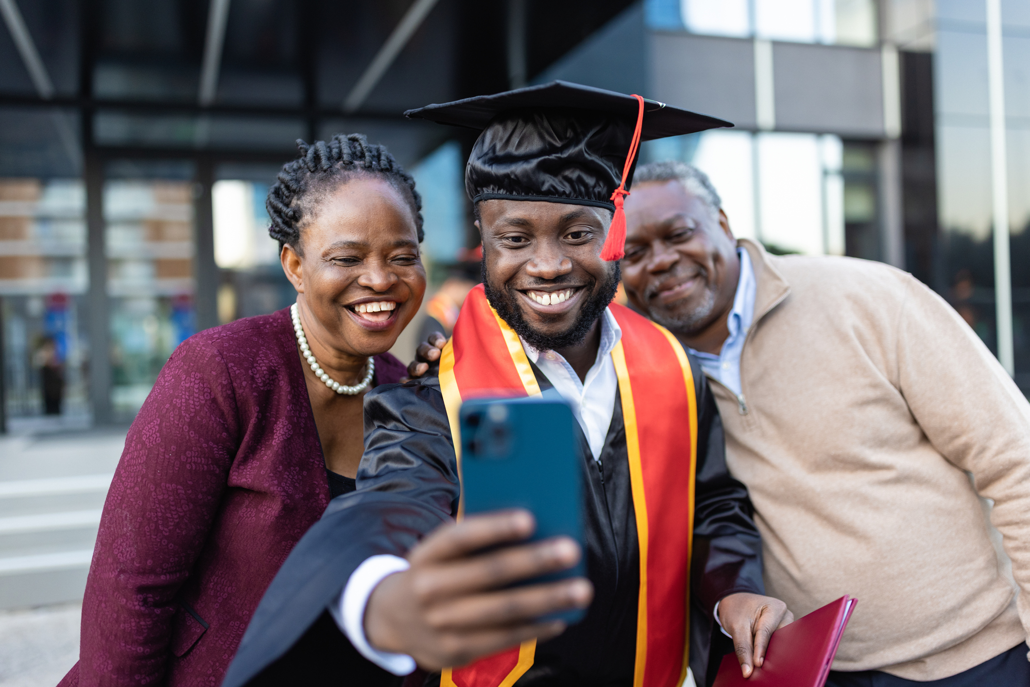 Someone in a graduation cap and gown taking a selfie with two people behind them.