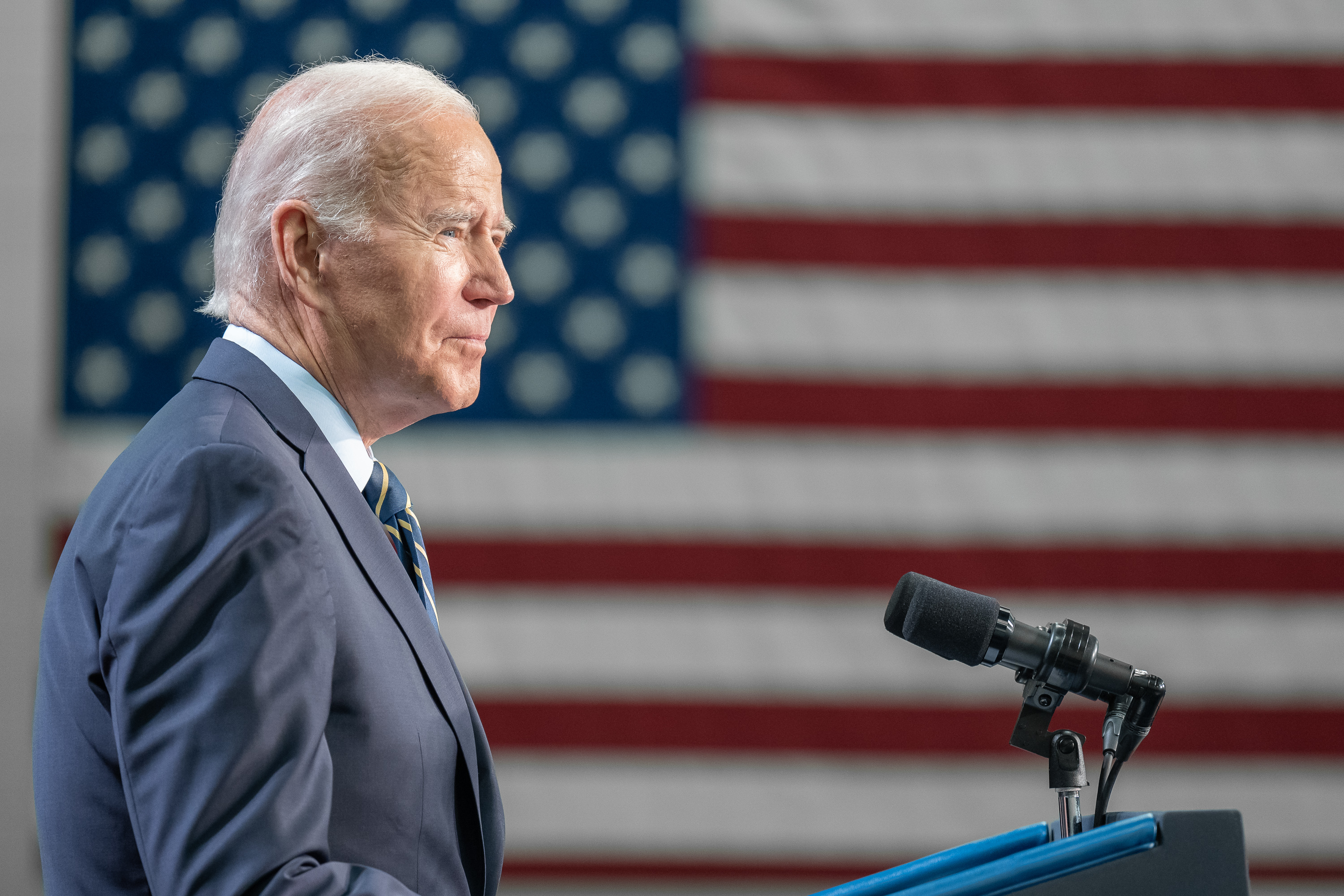 Joe Biden delivering remarks to a crowd, with a large American flag hung in the background.
