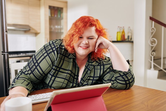 A person is smiling while reading an electronic tablet in a kitchen.