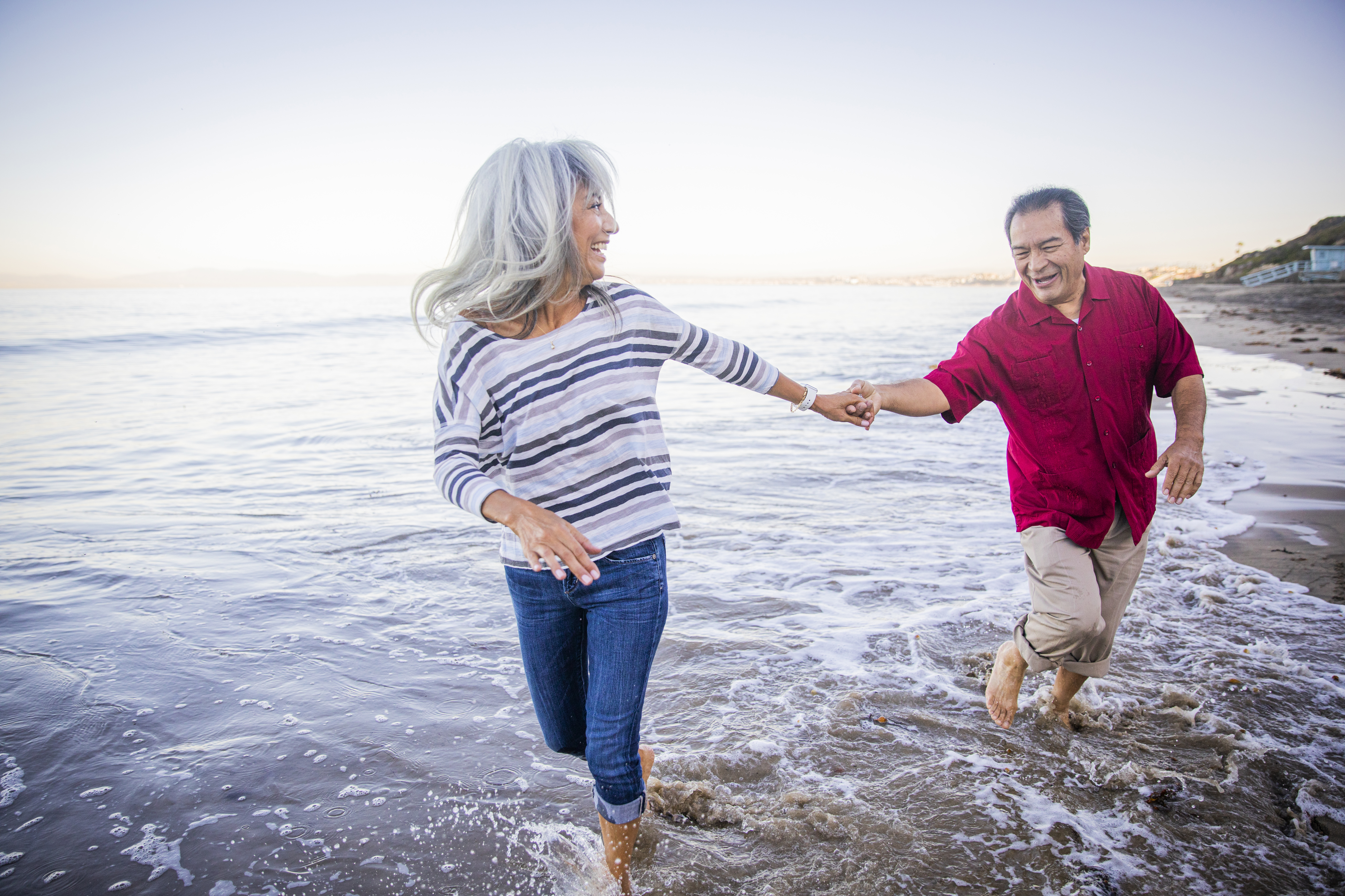 Two people holding hands and running on the beach through waves.