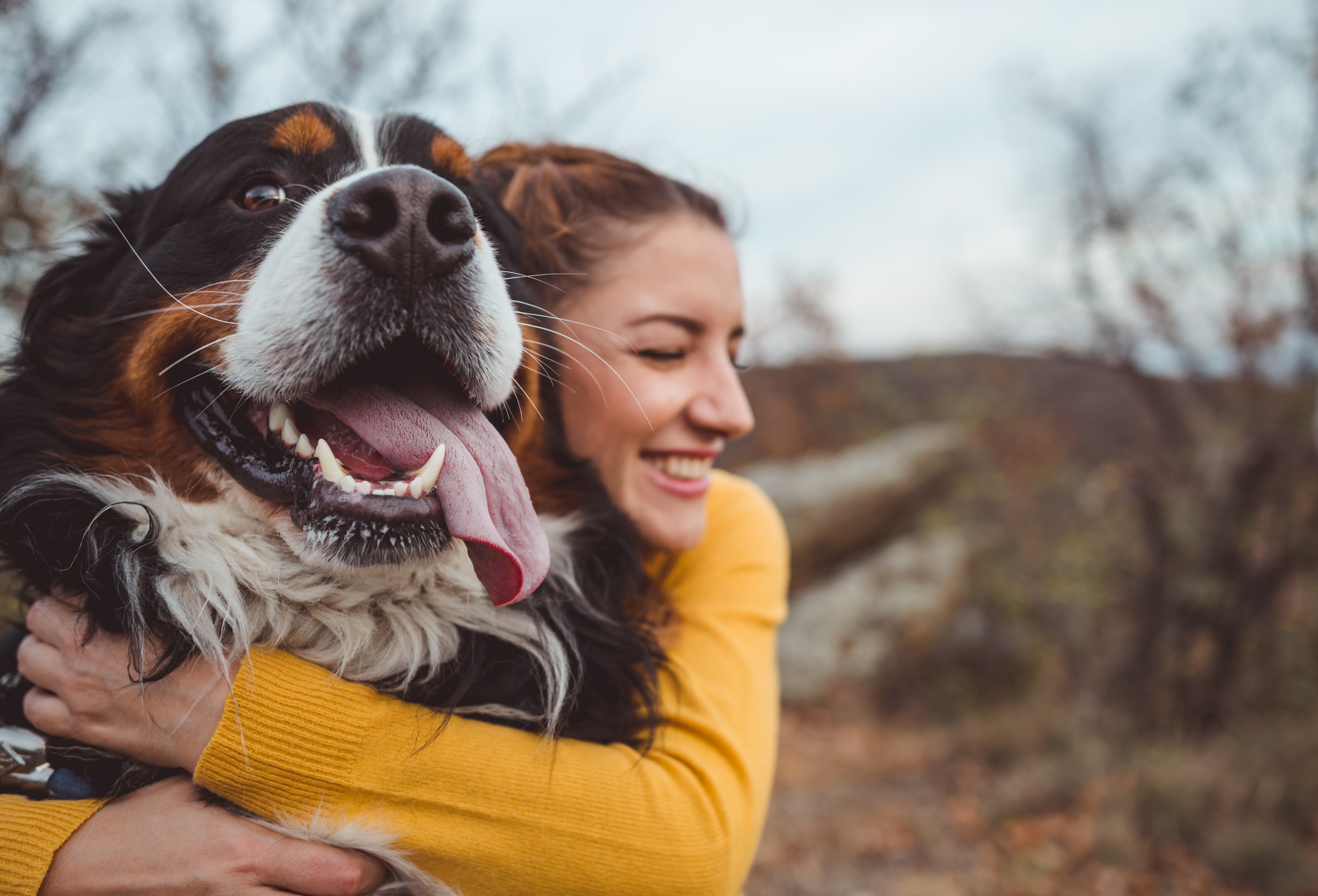 Someone is outdoors, hugging their dog and smiling.