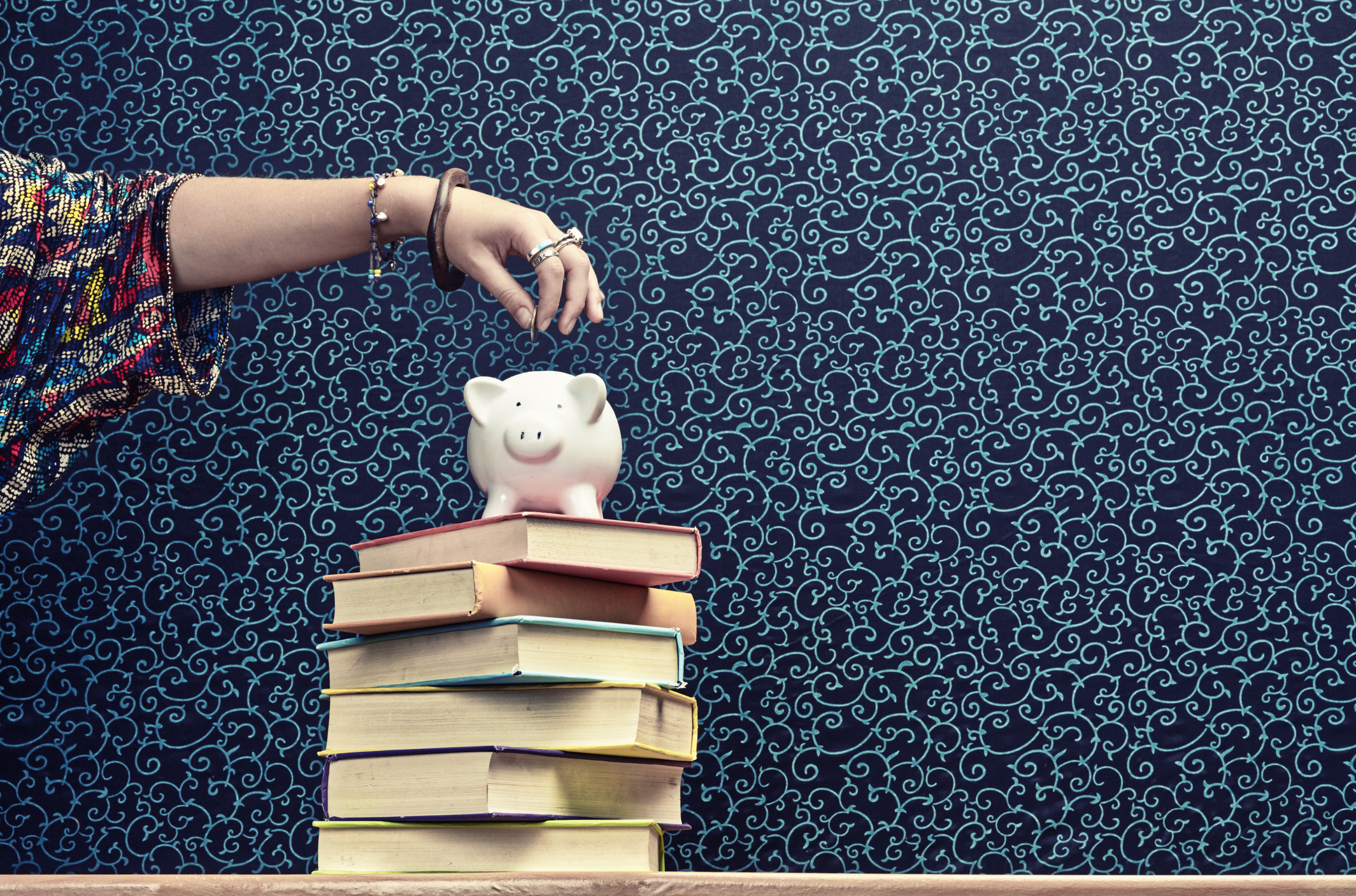 Hand putting coin in piggy bank atop a stack of books.
