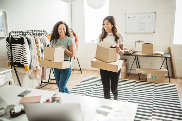 Two people in a boutique office hold shipping boxes.