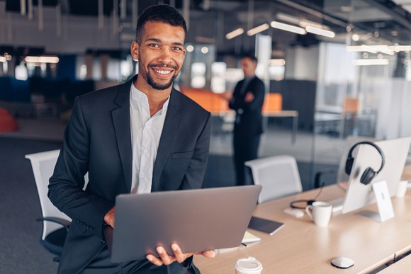 A smiling person in professional attire holding a laptop.