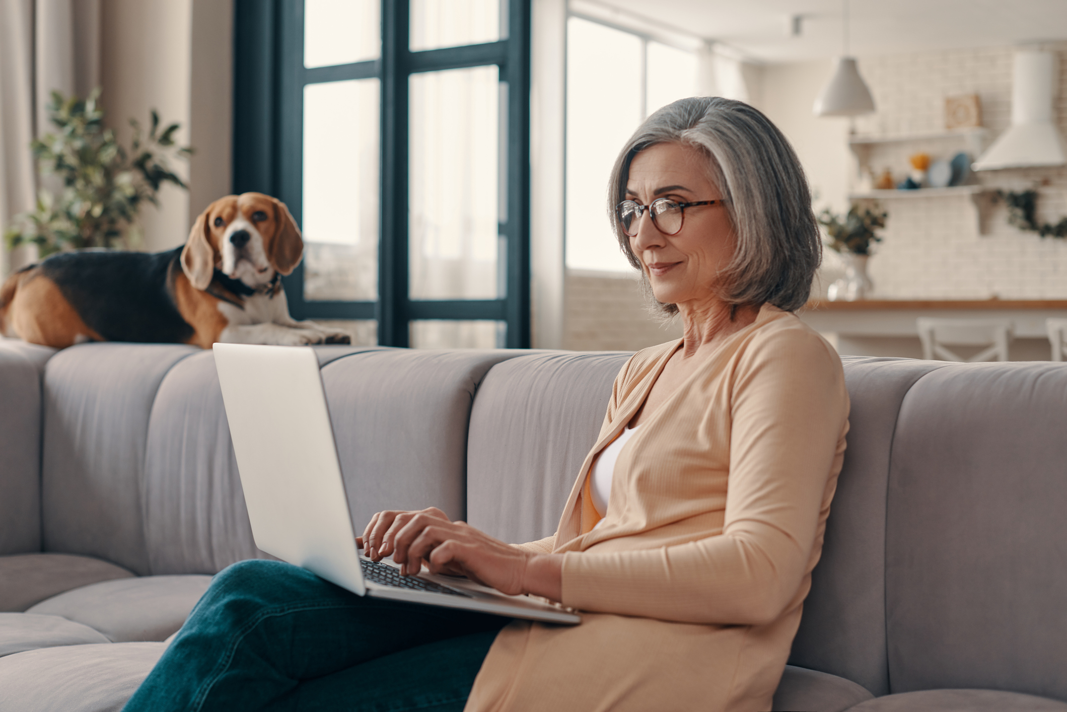 A person using a laptop on a couch with a dog looking on.
