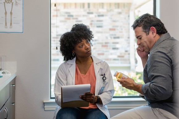 A doctor with a notepad sits with a patient who's holding a bottle of pills.