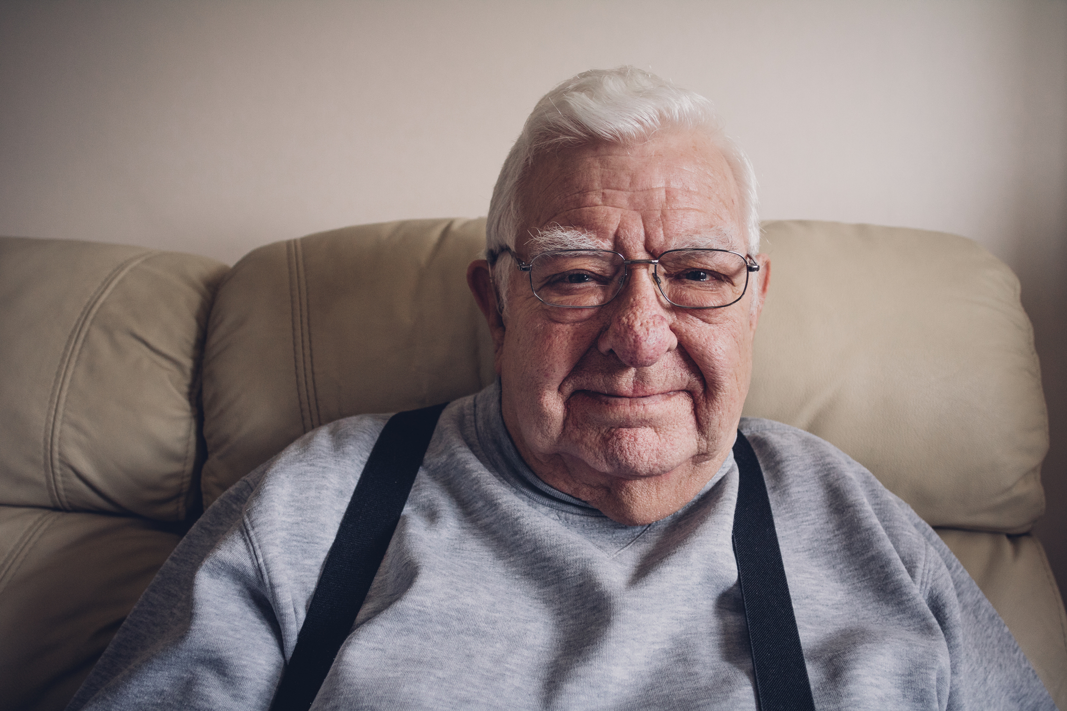 A senior citizen sitting on a couch, wearing suspenders, is smiling.