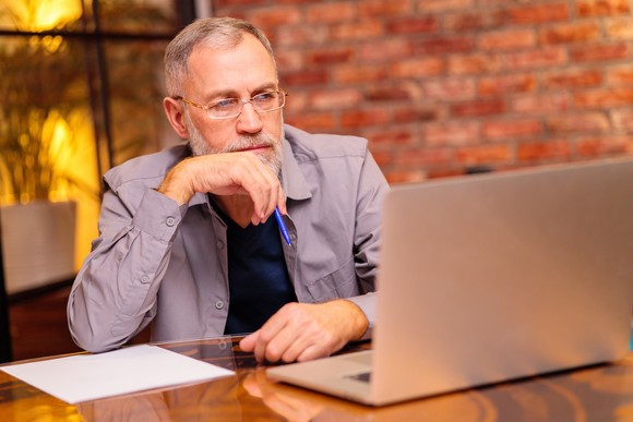 A person looking inquisitively at a computer screen while sitting at a table and holding a pen close to their mouth.