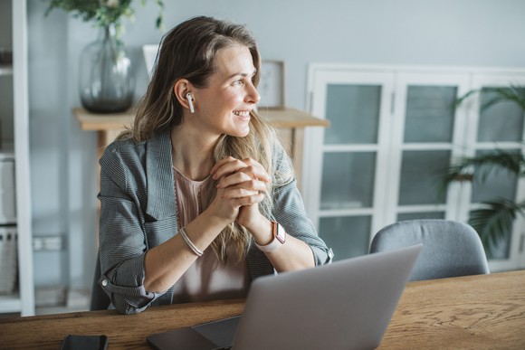 A person clasping their hands while sitting at a table with a laptop and looking to the side with a wireless earbud in their ear. 