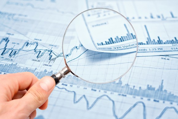 Hand holding magnifying glass above paperwork displaying stock charts and volume data.